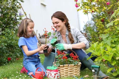 In a lush, well-maintained backyard garden near Kensington, a woman and a young girl are engaging in gardening activities amid green foliage, colorful flowering plants, and a neatly edged lawn. The woman, smiling and wearing green gardening gloves, is holding a small potted plant with red flowers, while the girl, dressed in a blue t-shirt and pink shoes, is giving her a watering can. The garden features a variety of plants, including a hedge on the right, a flowering shrub with pink blossoms in the background, and several other bushes and flowers contributing to the vibrant natural environment. A wicker basket with gardening tools is positioned in front of them, and a small, decorated watering can is placed on the grass nearby. The scene suggests a peaceful, outdoor space suitable for family gardening and landscape enhancement, reflecting the professional landscaping and garden renovation services offered by Landscapers Kensington in the local area. The lighting indicates a bright, partly cloudy day typical of spring or early summer, ideal for outdoor gardening work.