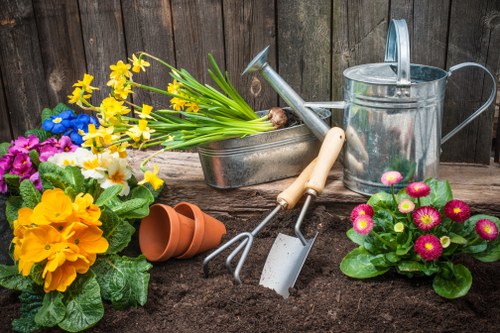 A scene of a well-maintained backyard garden in Kensington showing a freshly turned soil bed surrounded by vibrant flowering plants, including pink primulas and yellow primroses, with potted flowers arranged nearby. The garden features a wooden fence backdrop, and gardening tools such as a watering can, a hand rake, and a trowel are placed on the soil to suggest ongoing planting or maintenance work. In the foreground, there is an area of dark, rich soil prepared for new plantings, with a small terracotta pot lying on its side. The overall environment appears organized, with natural daylight illuminating the scene and a calm outdoor atmosphere suitable for gardening services offered by Landscapers Kensington in the Kensington area. The image emphasizes gardening activities, outdoor planting, and garden care, aligning with local landscaping and garden renovation services.