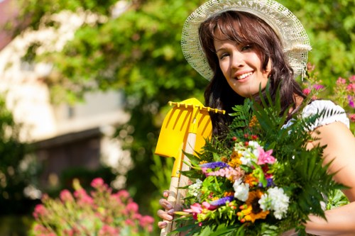 A woman standing in a well-maintained garden during daytime, holding a yellow watering can and a large bouquet of colorful flowers, with lush green hedges, trees, and flower beds in the background. The garden features a neatly trimmed lawn, flower borders with vibrant blooms, and a paved pathway. The scene is sunny with clear weather, highlighting the natural tones of the garden elements. The outdoor space appears spacious and landscaped, suitable for gardening and outdoor maintenance services offered by local landscapers in Kensington.
