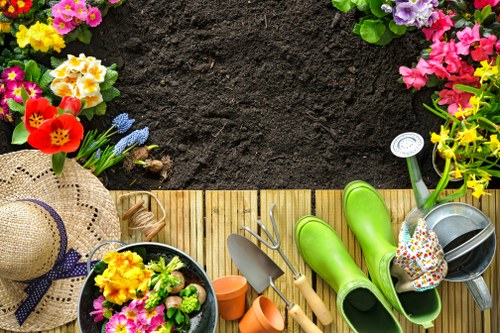 The image shows an organized outdoor garden space with a wooden deck adjacent to a freshly turned soil bed, prepared for planting. The foreground features a variety of gardening tools and accessories, including green rubber boots, a metal watering can, small terracotta pots, a gardening trowel, and a hand rake, all arranged neatly on the deck. To the left, there is a straw sunhat with a purple ribbon, alongside a black planter filled with blooming flowers in shades of yellow, pink, and purple, creating a vibrant contrast against the dark soil background. The garden borders are lined with colorful flowering plants and lush green foliage, suggesting a well-maintained landscaped yard typical of residential properties in the Kensington area. The scene is lit by natural daylight, indicating a clear, calm weather condition ideal for gardening. This outdoor space highlights a tidy, cultivated garden environment that exemplifies professional gardening and landscaping services offered by companies like Landscapers Kensington in London, supporting local gardening and lawn care needs.