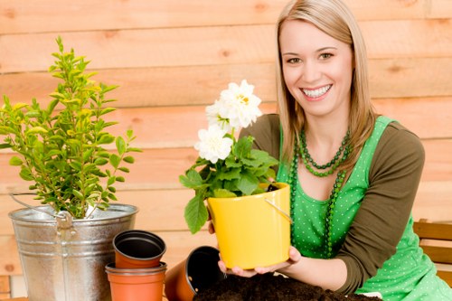 The image shows a smiling woman with blonde hair wearing a green dress with polka dots, sitting outdoors in a garden setting with a wooden fence in the background. In front of her, there is a bright yellow flower pot containing a blooming white flowering plant. To her left, there are various gardening containers, including a metallic bucket with a small green shrub, an empty black plastic pot, and a reddish-brown terracotta pot. The garden features a well-maintained grassy lawn in the foreground with dense, green grass, and adjacent flower beds or borders that may contain other plants or soil, contributing to a lush, verdant environment. The overall scene suggests an active gardening space, ideal for landscaping or outdoor maintenance, with natural daylight highlighting the vibrant colours and textures of the plants, containers, and wooden fencing, aligning with professional gardening and landscaping services in Kensington and surrounding areas.